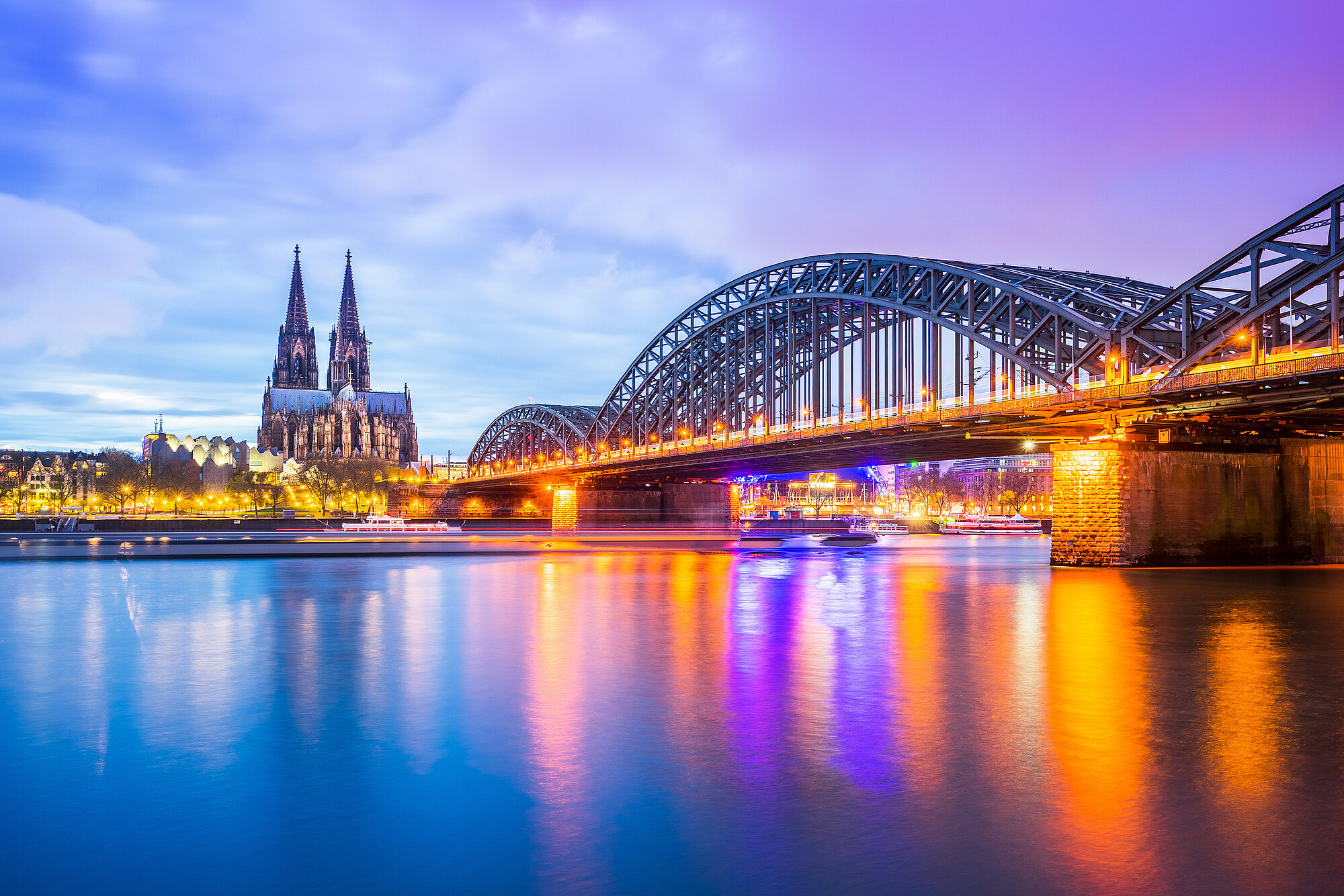 Skyline von Köln bei Dämmerung, mit dem Rhein und der Hohenzollernbrücke im Vordergrund und dem Kölner Dom im Hintergrund