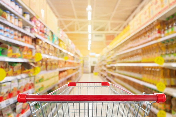 Blick auf einen Einkaufswagen und auf einen Gang in einem Supermarkt mit Lebensmitteln