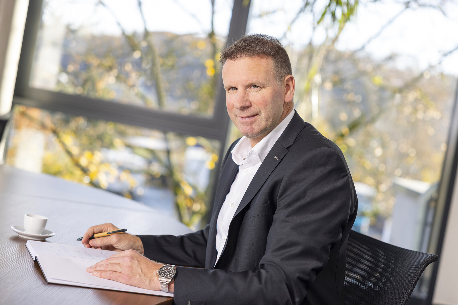  Ralf Siefen in the office, sitting in front of a book and a coffee cup, wearing a suit with a wristwatch and pen in hand, starts to write