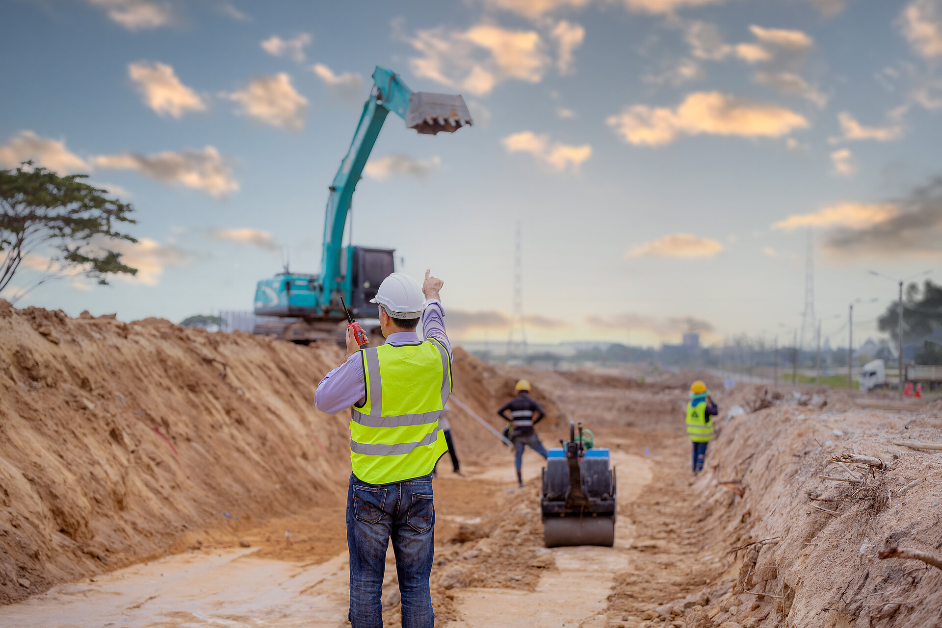 Mann in Warnweste steht vor einer Baustelle mit Bagger neben einer Grube, in der eine weitere Baggerschaufel liegt; zwei weitere Männer mit Warnwesten und Helmen stehen ebenfalls vor Ort.