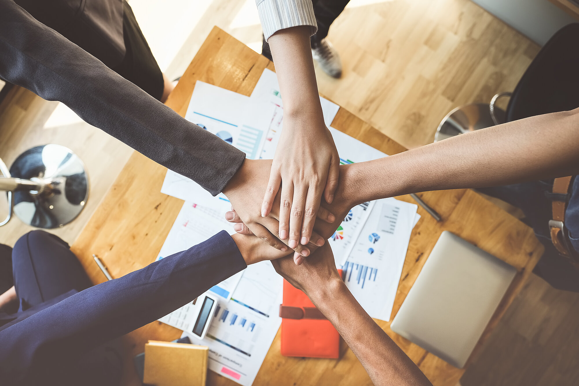 5 Personen, die über einem Schreibtisch als Zeichen des Teamworks jeweils eine Hand aufeinander legen