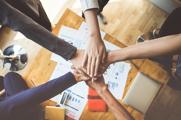 5 Personen, die über einem Schreibtisch als Zeichen des Teamworks jeweils eine Hand aufeinander legen