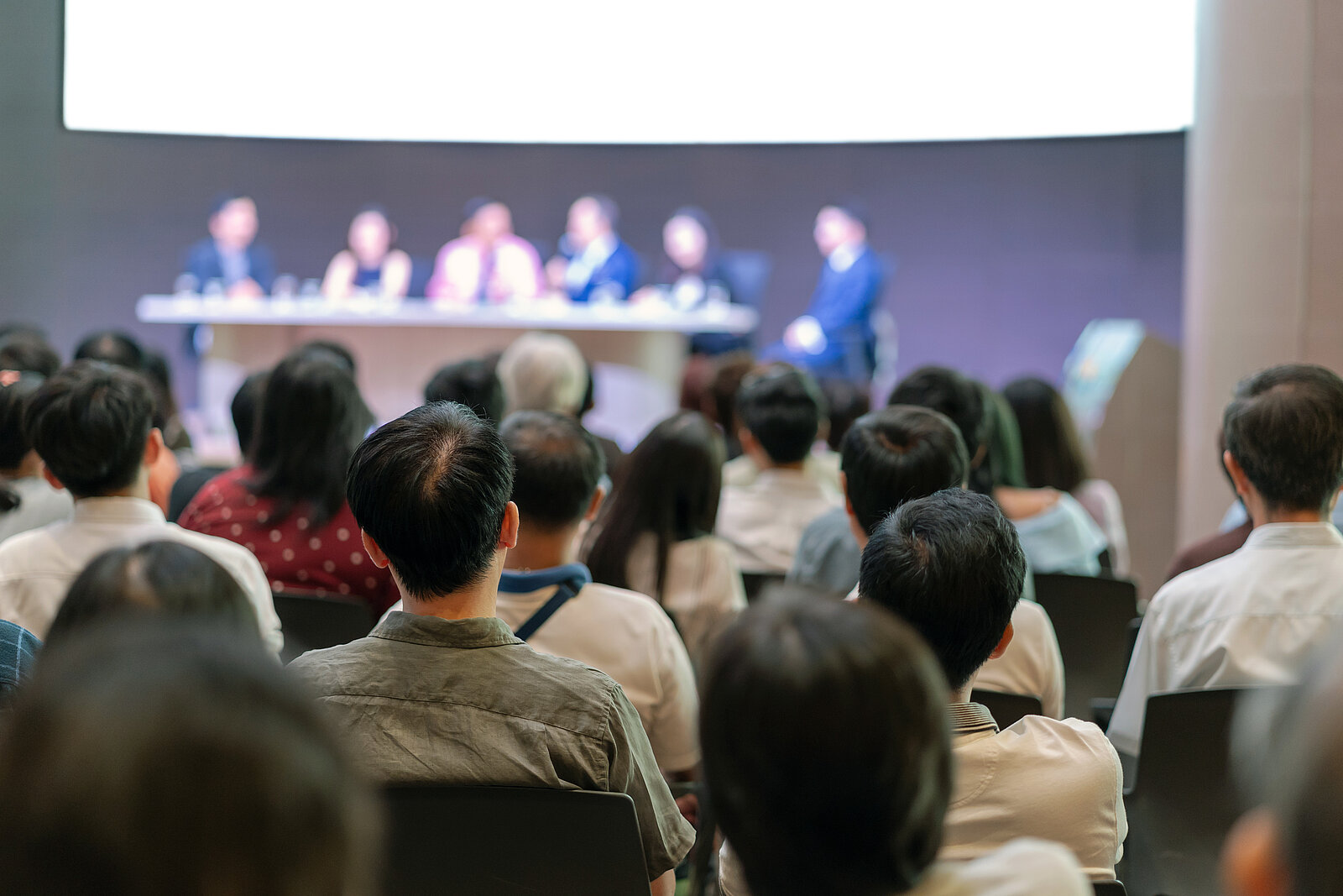 Spectators watch a discussion between six people sitting at a table on a stage.