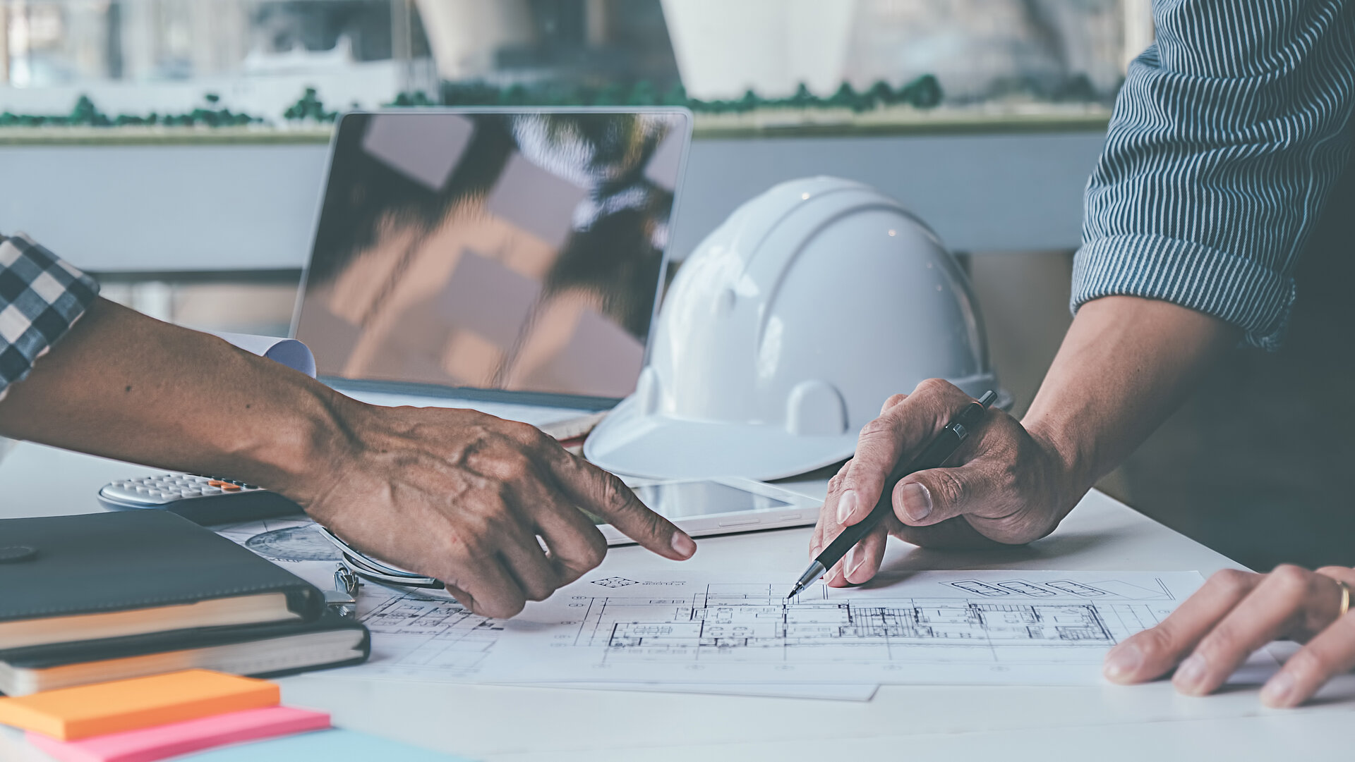 Architekt und Ingenieur stehen gemeinsam an einem Schreibtisch und planen einen Bauentwurf, im Hintergrund liegen Laptop und Bauhelm auf dem Tisch.
