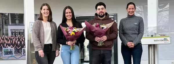 The trainer and HR manager flank the two training graduates, each holding a bouquet of flowers. They are all standing and smiling in front of a carpet with the words “Welcome” written on it.