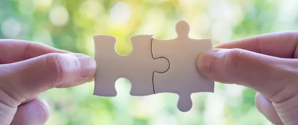 2 hands, each holding a puzzle piece with thumb and index finger, which fit into each other