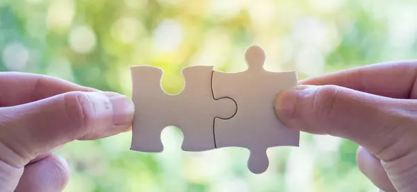 2 Hände, die jeweils mit Daumen und Zeigefinger ein Puzzleteil festhalten, welche ineinander passen