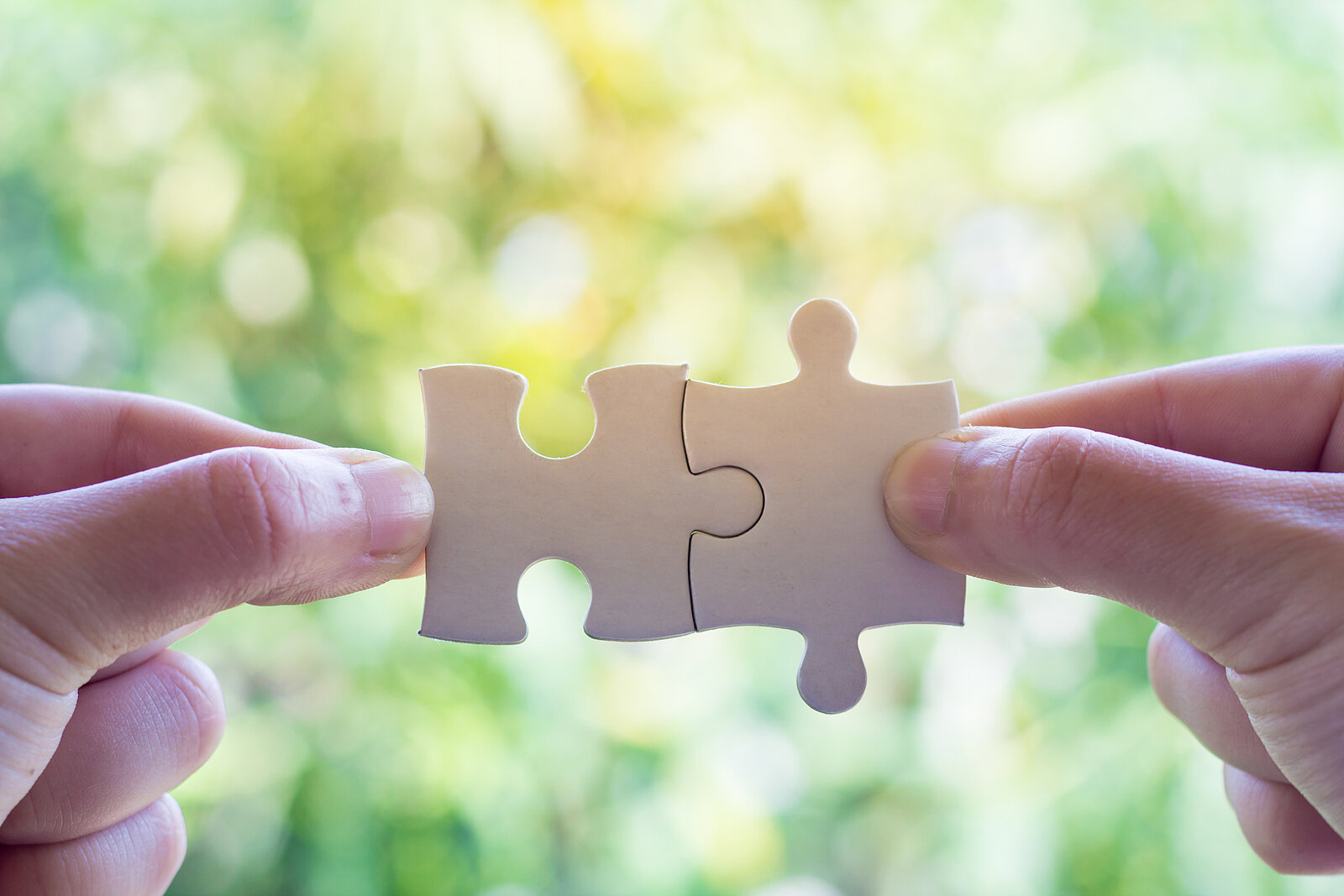 2 Hände, die jeweils mit Daumen und Zeigefinger ein Puzzleteil festhalten, welche ineinander passen