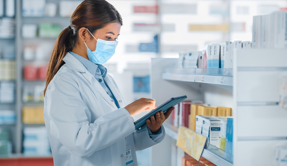 Eine Frau mit Mundschutz in einer Apotheke arbeitet an einem Tablet. Im Hintergrund ein Schrank voller Medikamente