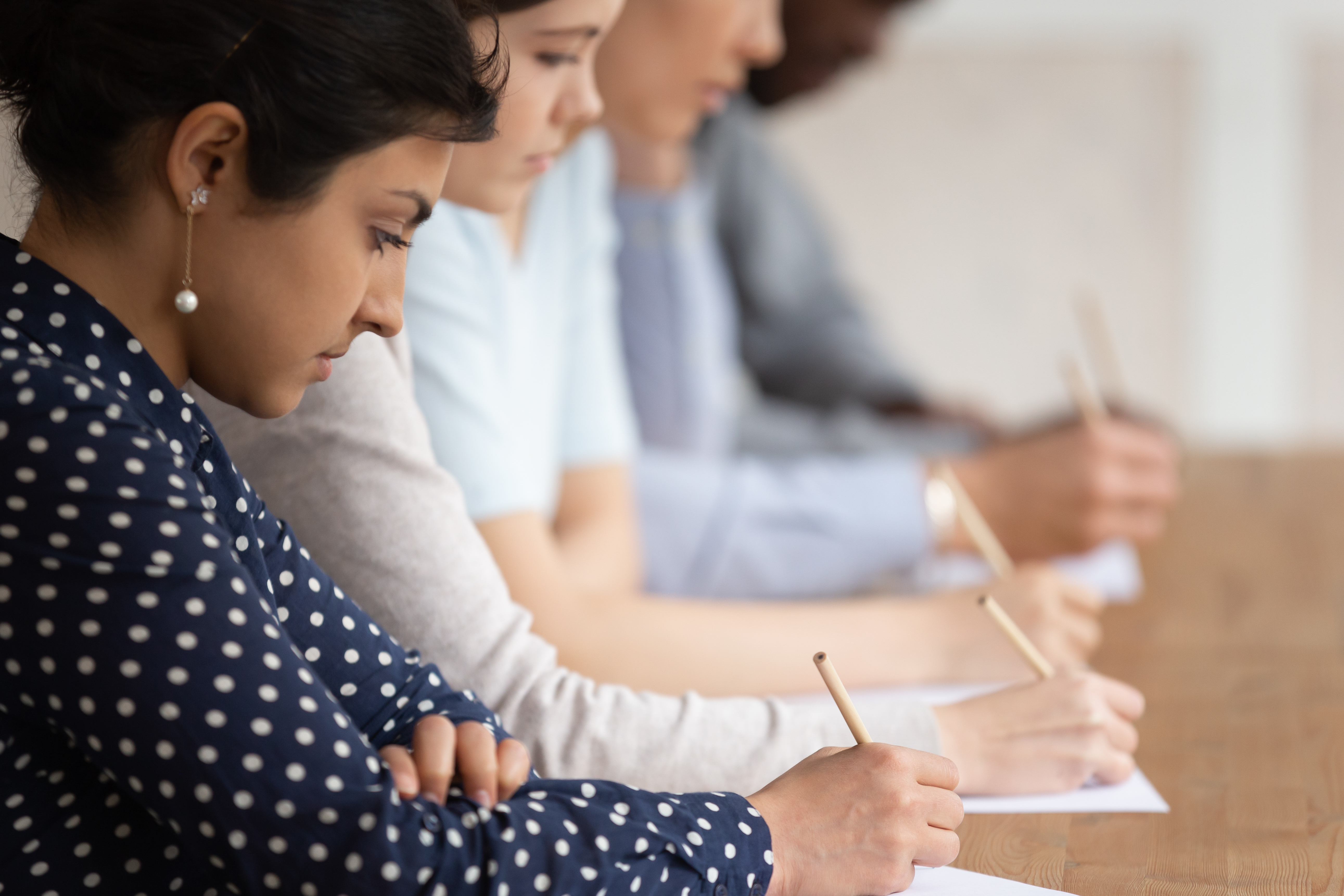 Students sit in a row at a desk and write with pencils on paper.