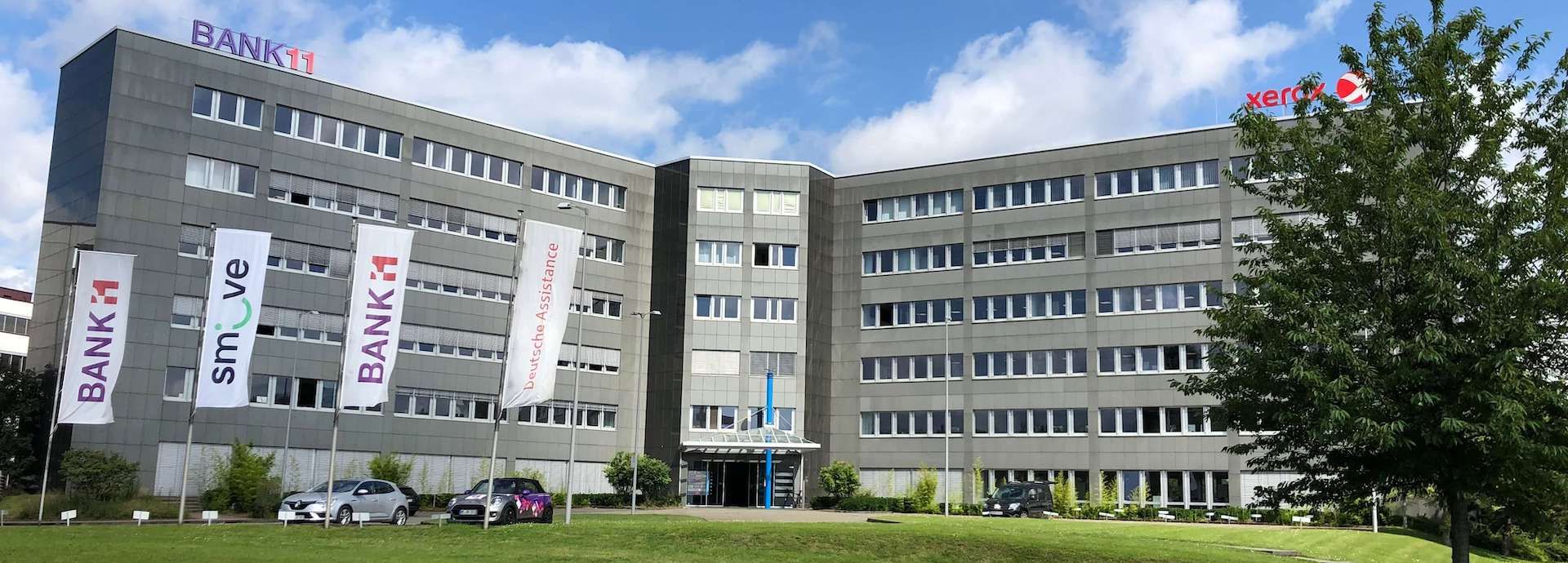 Das Gebäude von Bank 11 mit 4 Fahnen, einer Grünfläche und einem Baum davor