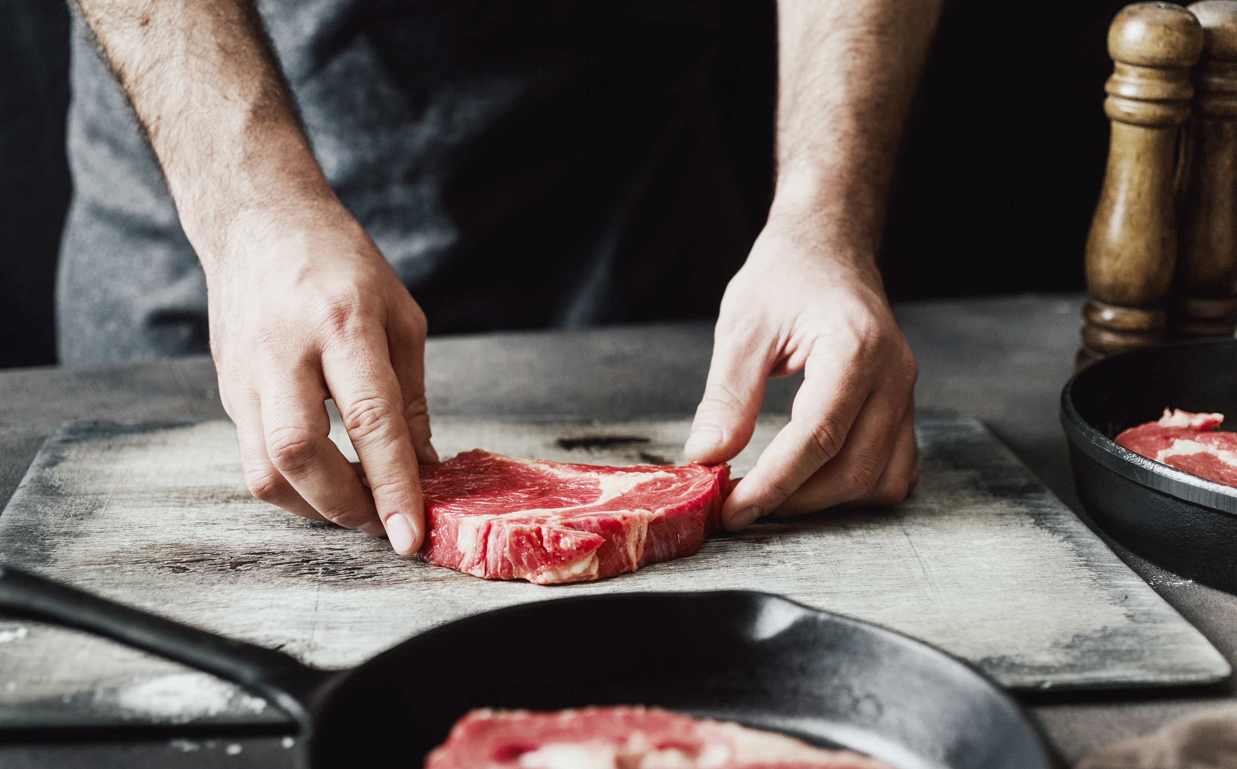 Mann verarbeitet ein Stück Fleisch, im Vordergrund eine Pfanne, rechts ein Pfefferstreuer