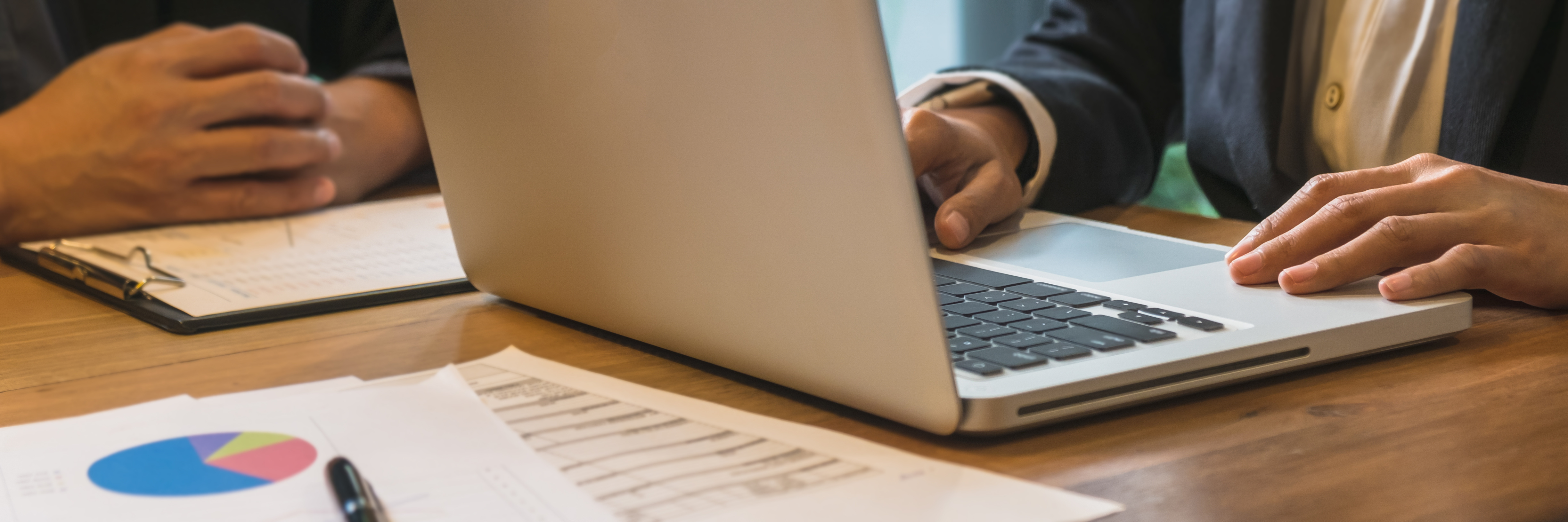 2 Menschen sitzen an einem Schreibtisch, einer arbeitet an einem Laptop, der andere hat die Hände ineinander geschlagen und hat Papier vor sich